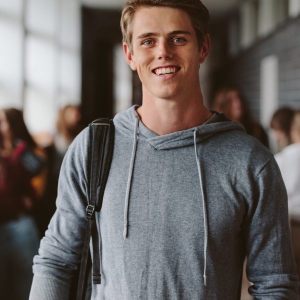 Portrait of happy young guy with bag walking in university corridor with classmates in background. Young male student in college campus.